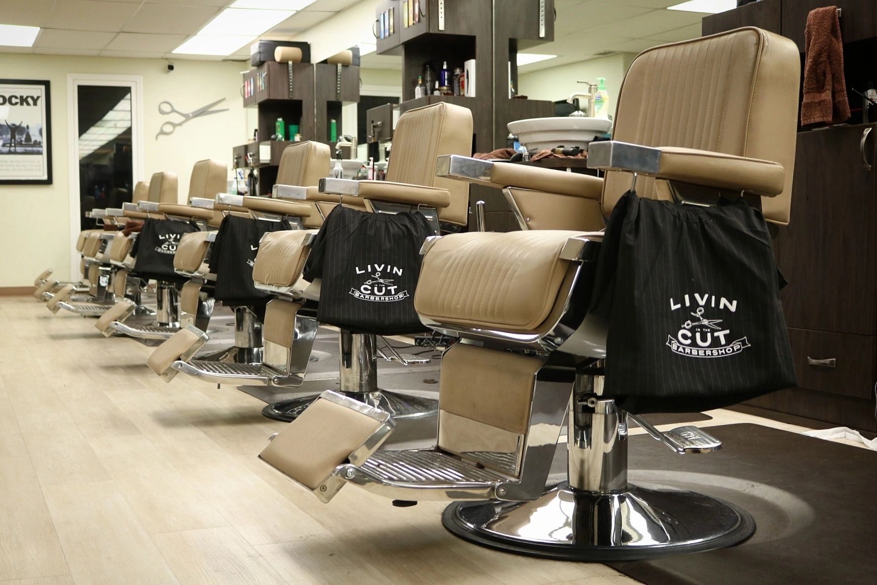 A row of barber chairs at Livin In The Cut, each with a pinstriped cape over the back.